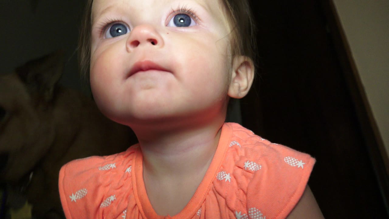 A toddler girl in a coral patterned top looks upward from a low floor-level angle inside a Broomfield, Colorado home.
