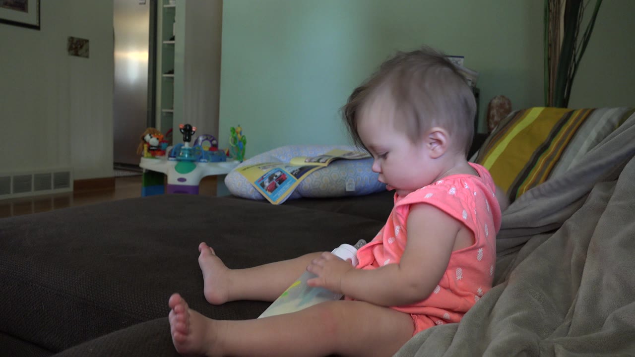 Toddler in a pink outfit sitting on a dark couch with a bottle in her lap.