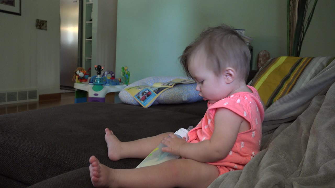 The toddler remains seated in profile in the Broomfield home, looking down while holding or steadying a baby bottle against the dark couch surface.