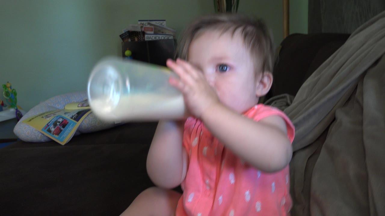 A baby in a pink outfit drinks from a translucent bottle on a dark couch inside a Broomfield, Colorado home, with a green wall and household items behind her.