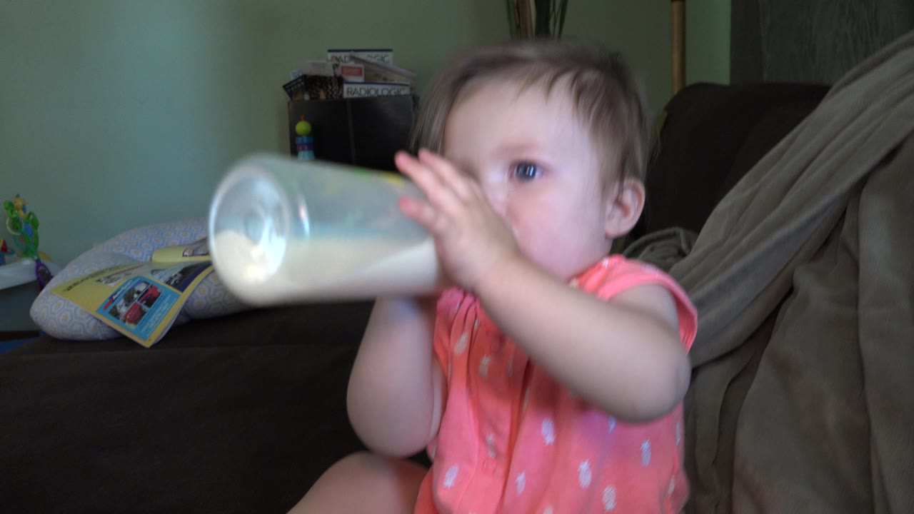 The baby keeps both hands on the bottle while seated on the couch in Broomfield, Colorado, with pillows and a partially visible household shelf in the background.