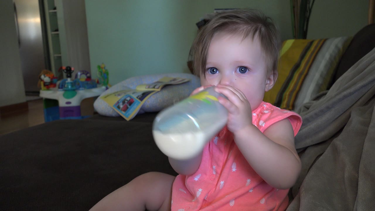 A toddler in Broomfield, Colorado sits on a dark bed and drinks from a translucent bottle, with pillows and children's toys visible behind her.