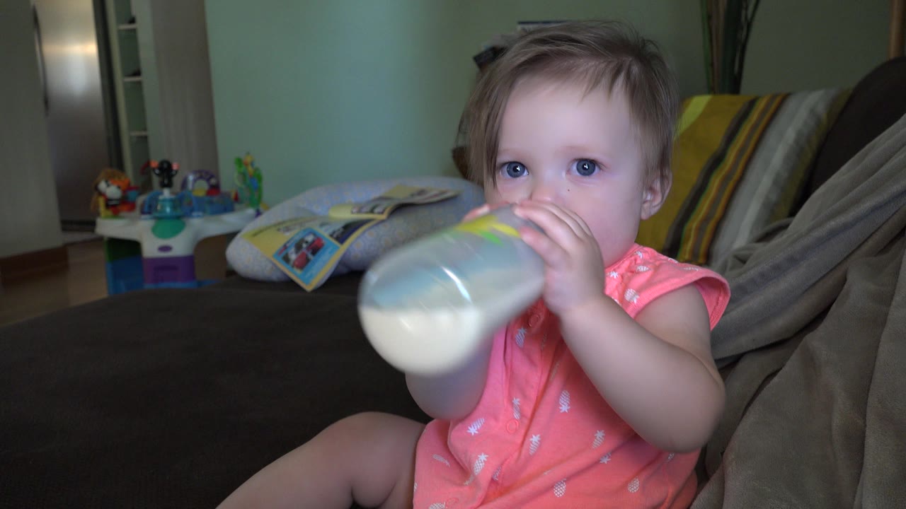 Toddler holds a bottle to her mouth on a bed.