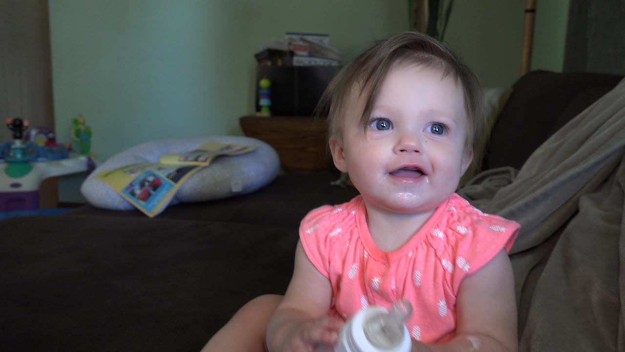 At 0:01 in Broomfield, Colorado, the child smiles slightly while seated on the couch, with the bottle low in the foreground and home toys visible in back.