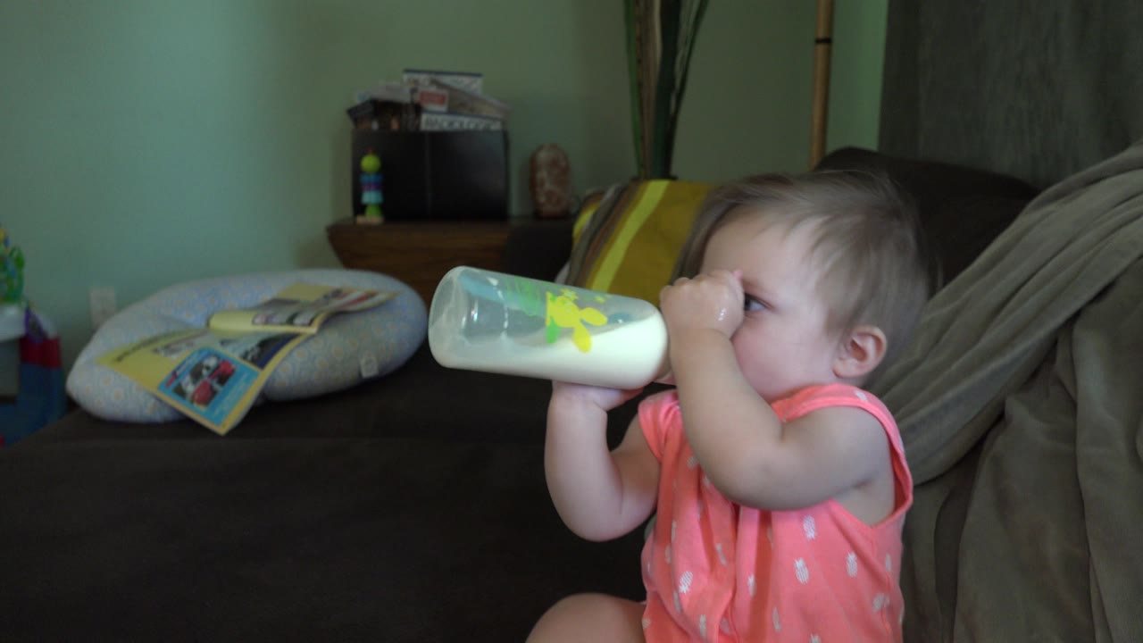 A baby in a coral outfit drinks from a translucent formula bottle on a dark couch in a Broomfield, Colorado living room.