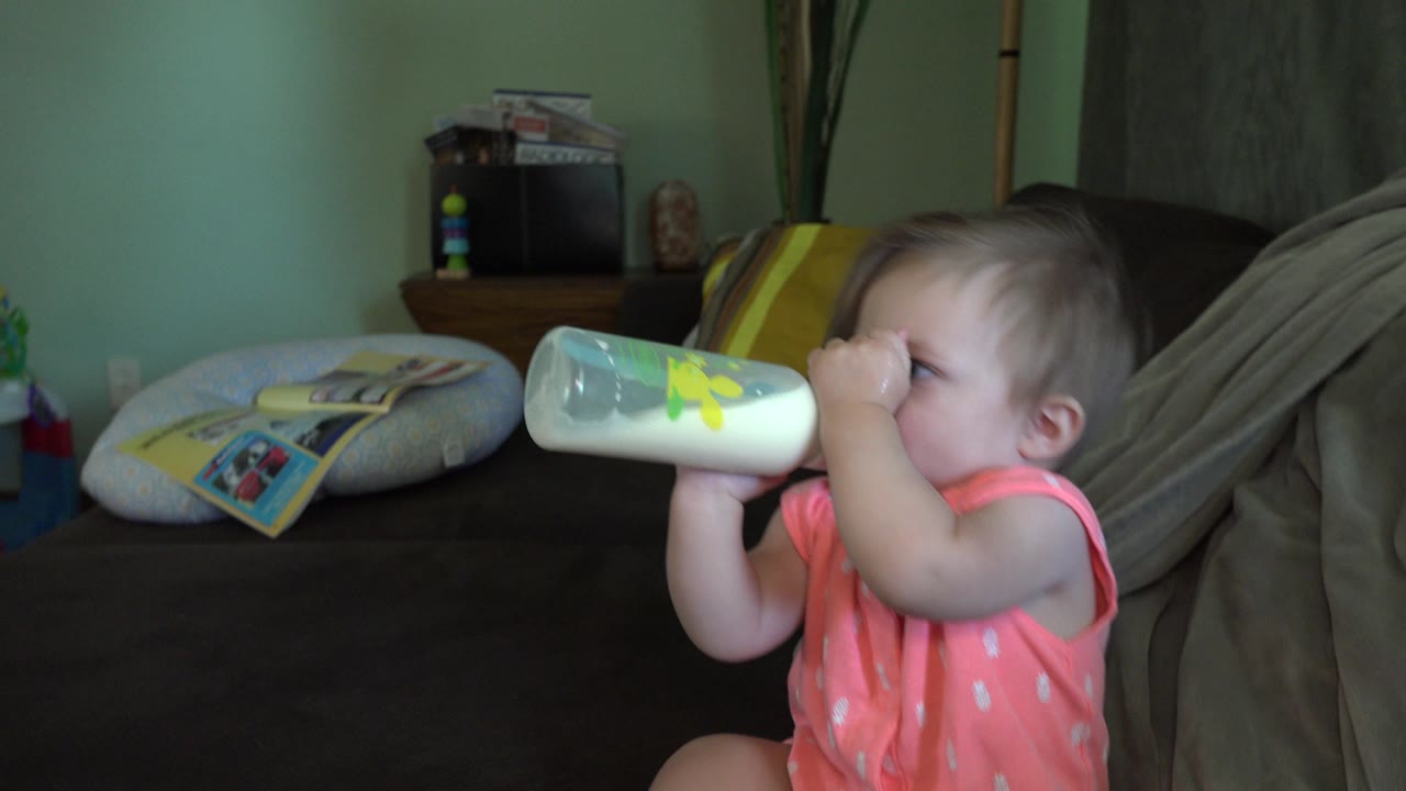 The baby keeps the bottle lifted to the mouth while facing right, with couch cushions and a side table behind in the Broomfield home.
