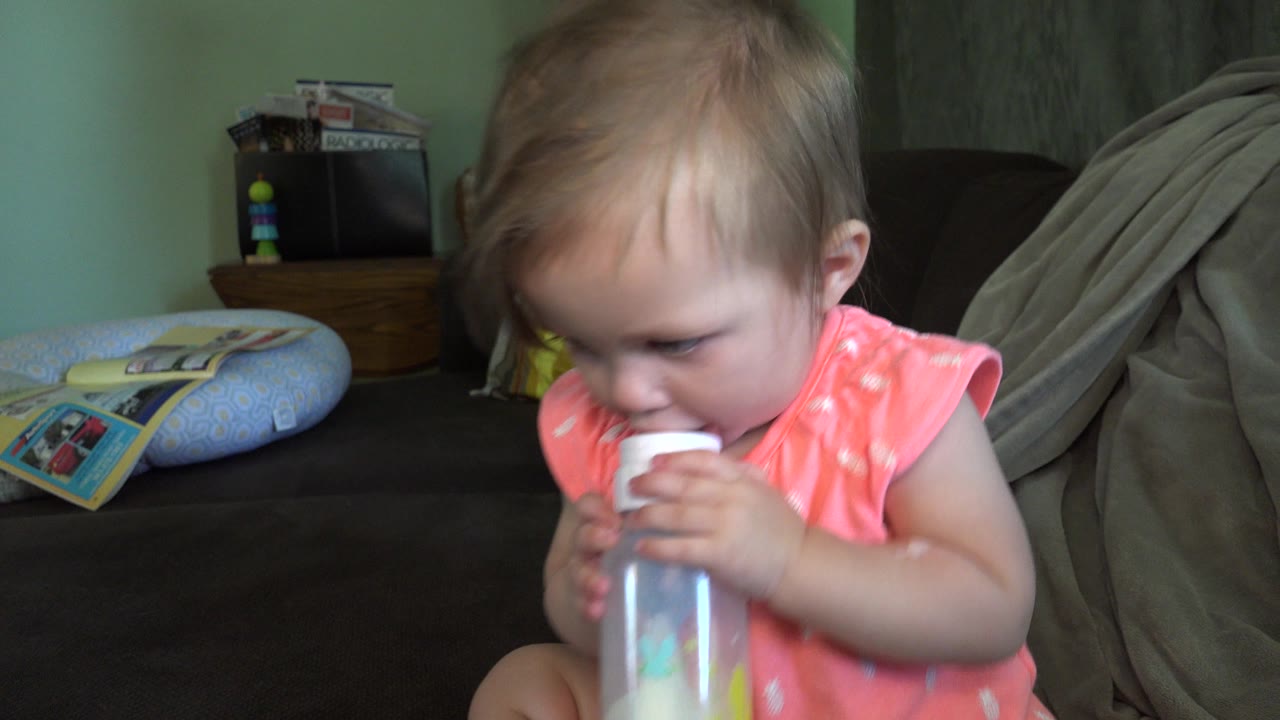 Baby girl in a pink outfit drinking from a bottle on a couch.
