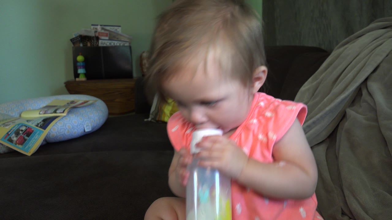 The baby remains close to the camera with the bottle at her mouth, while a pillow and blanket sit on the dark couch behind her in the Broomfield home.