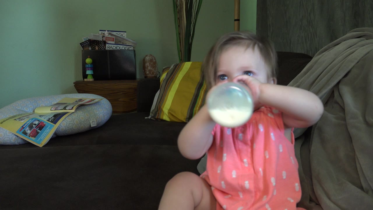 A baby in a pink outfit drinks from a translucent bottle on a dark couch inside a Broomfield, Colorado home, with striped pillows and a side table behind her.