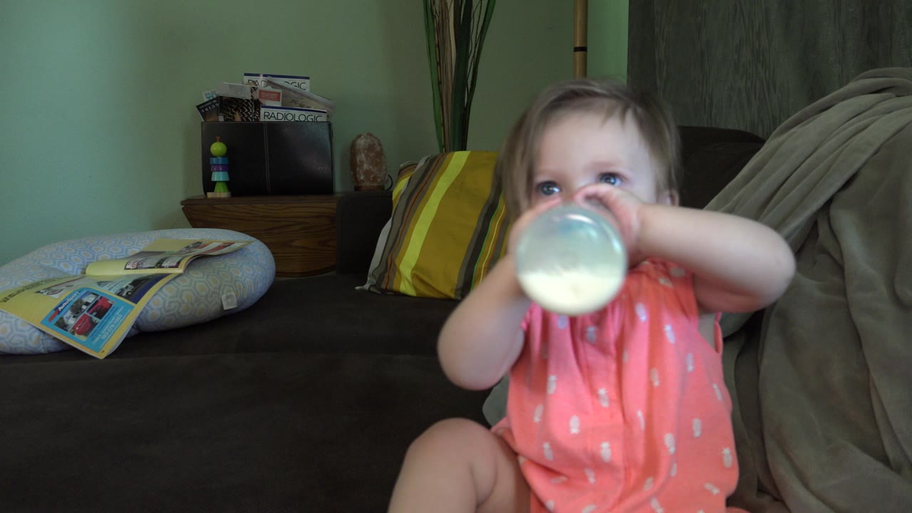 The baby continues drinking from the bottle on the couch, with the bottle nearer to the lens and the Broomfield home interior still visible in soft indoor light.