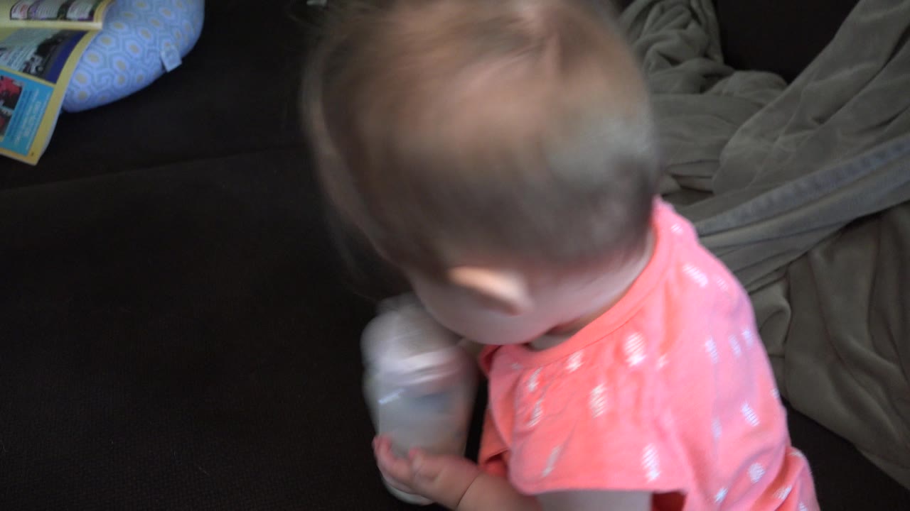 In Broomfield, Colorado, a blond baby girl in a coral pink outfit looks down while gripping a clear bottle near a dark couch and gray blanket.