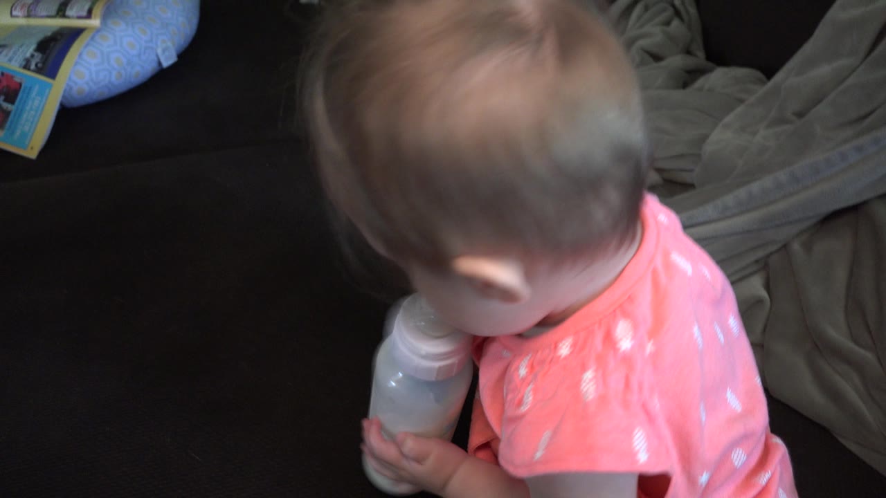 The Broomfield home clip shows the baby girl with her head lowered toward the bottle, with the dark couch and rumpled gray blanket filling the background.