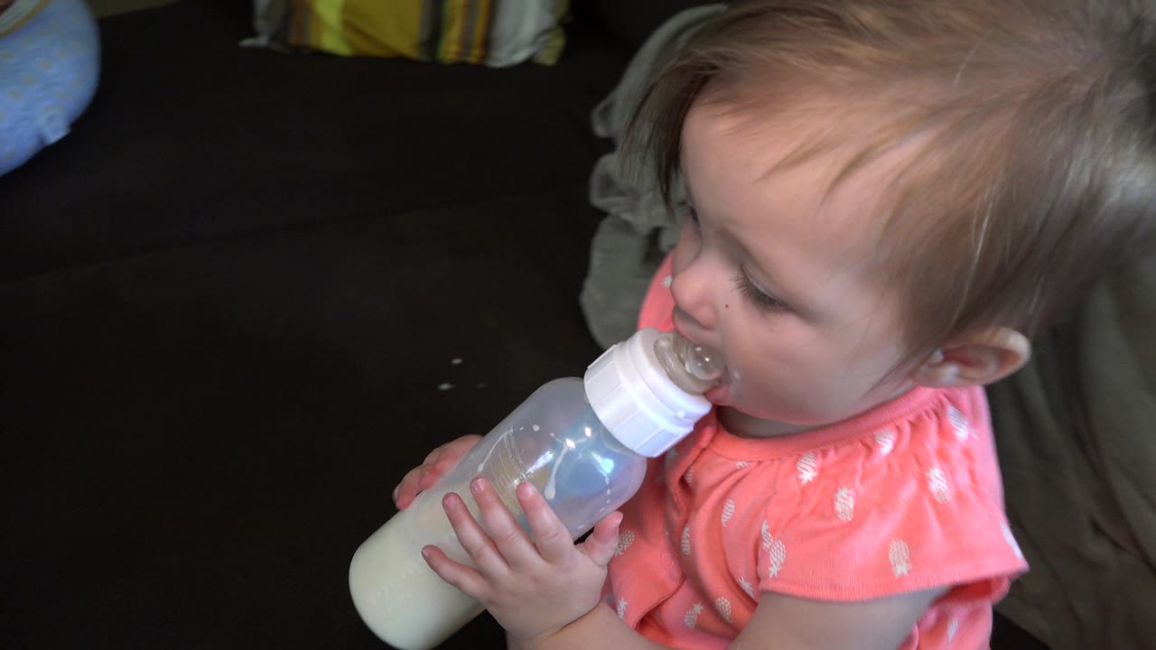 In the Broomfield, Colorado interior, the baby girl drinks from a clear bottle in side profile, with pale milk visible inside and the couch behind her.