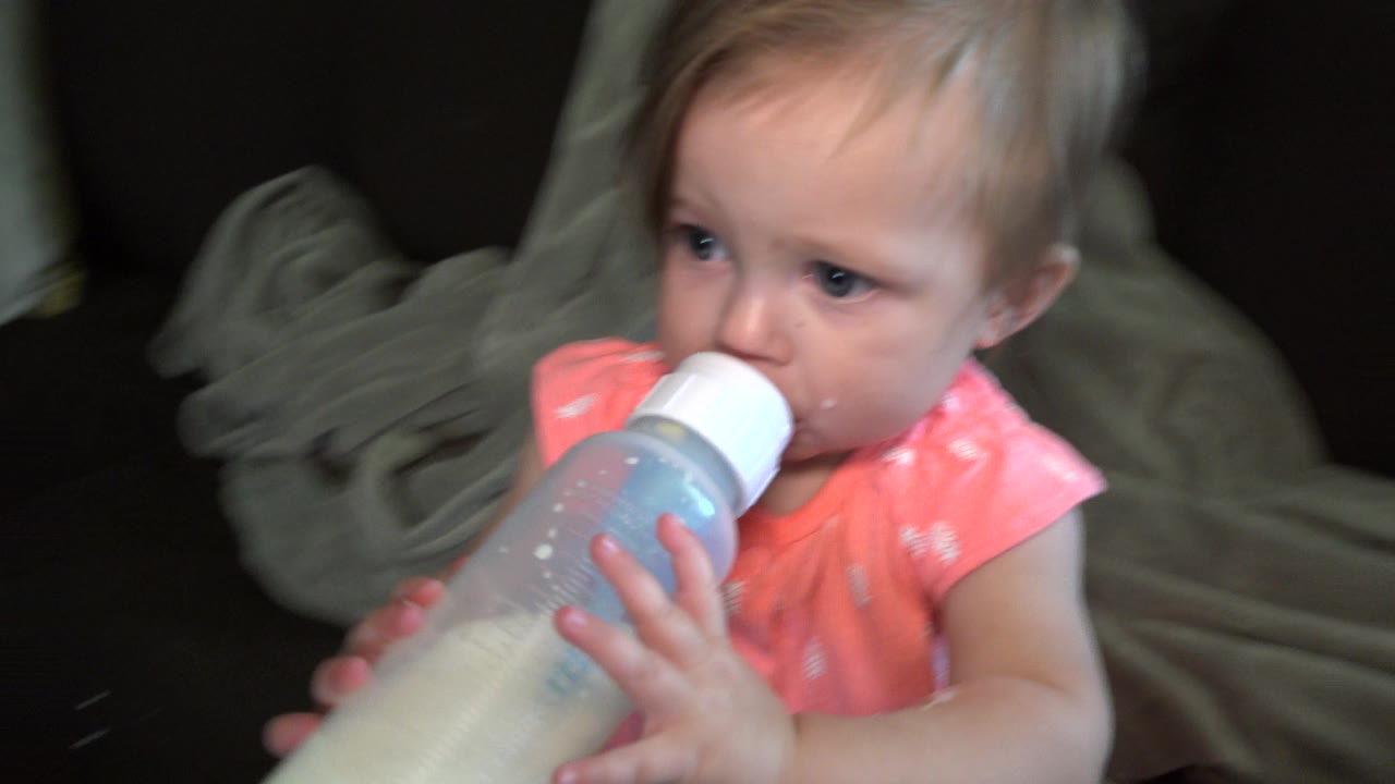 Baby girl drinks from a bottle while facing left on the couch.