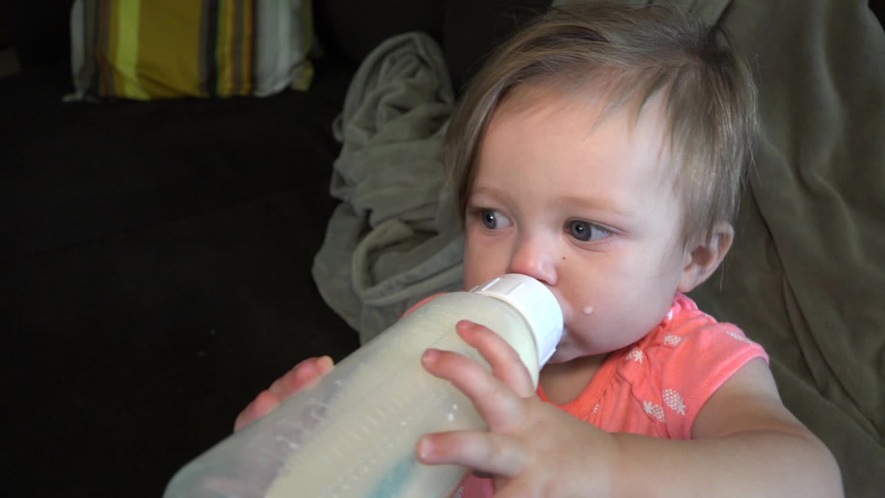 A toddler in a coral polka dot shirt drinks from a large milk bottle on a dark sofa in Broomfield, Colorado.