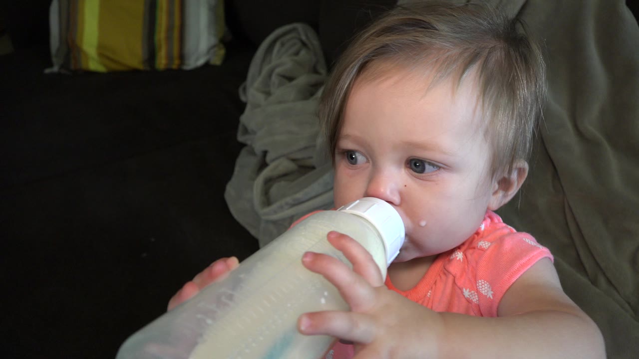 The child continues drinking from the bottle while looking left, with soft light catching her face in the Broomfield home interior.