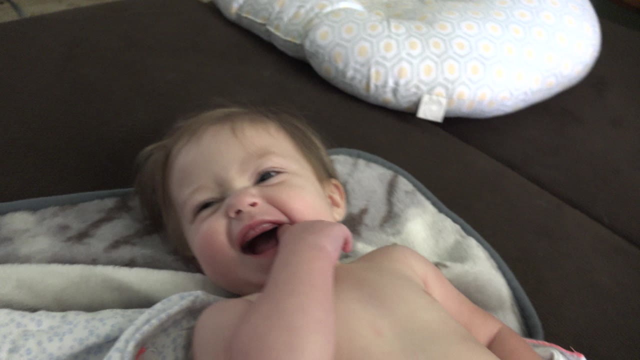 The baby continues smiling on the blanket in Westminster, Colorado, with the hand pressed closer to the mouth and the couch behind.