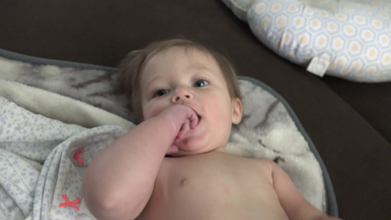 The infant keeps fingers in the mouth while lying on a light patterned blanket, with a pale pillow visible above the head in the Westminster home scene.
