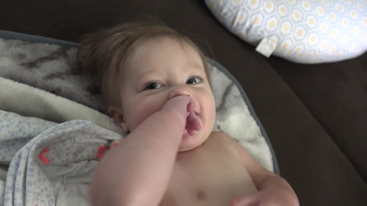 The toddler remains reclined on blankets in Westminster, Colorado, looking toward the camera with fingers at the mouth and pillows behind.