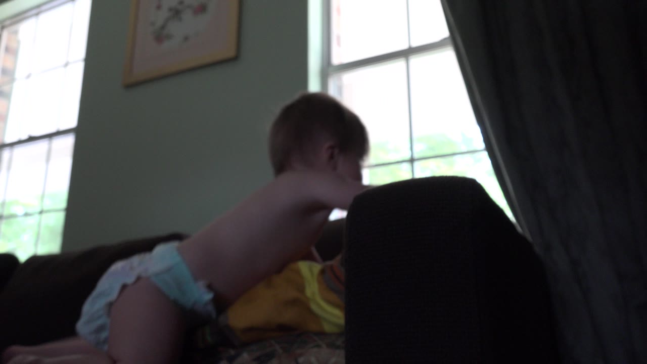A diapered baby crawls across dark furniture inside a Westminster, Colorado home, with a bright multi-pane window and dark curtain behind them.