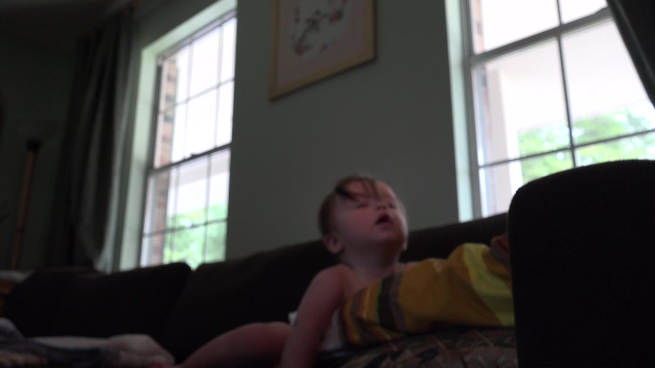 Baby reclines on a dark couch near bright windows.
