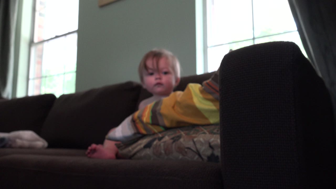 A baby girl sits on a dark sofa in a Westminster, Colorado home, partly covered by a striped blanket with bright windows and curtains behind her.