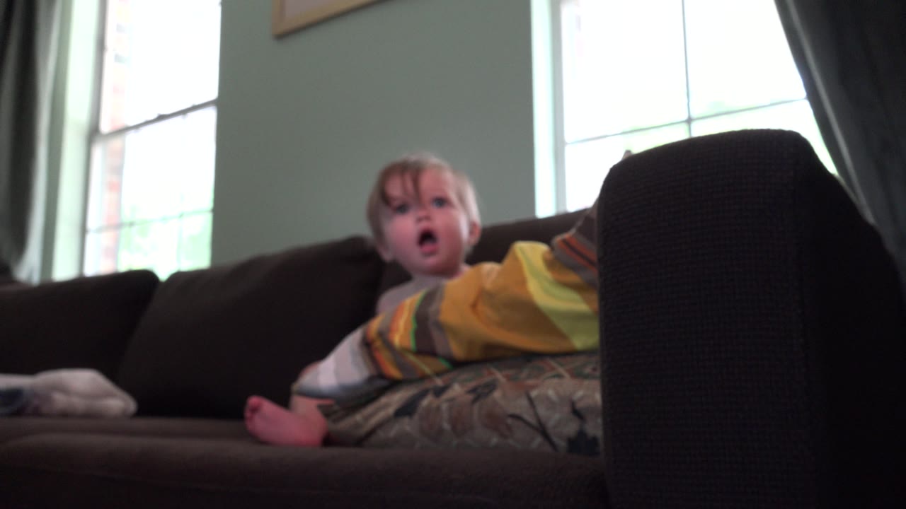 Baby girl on sofa with mouth open near bright windows.