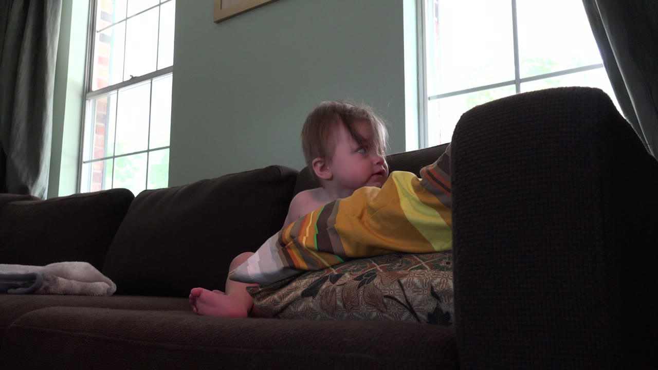 Baby boy seated on a brown sofa near colorful pillows and a bright window.