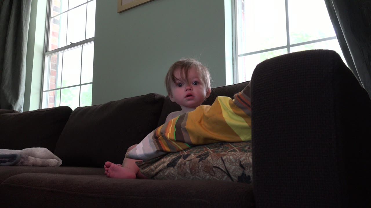Toddler sitting on a dark couch near a bright window.