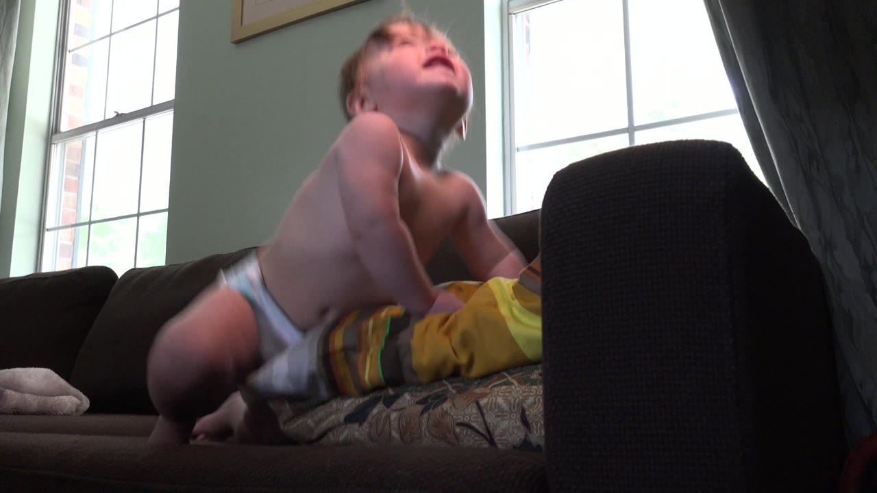 Baby in a diaper leaning on a sofa cushion near a bright window.