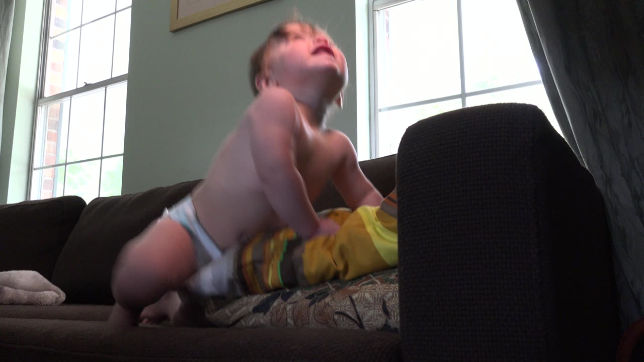 The baby arches their head upward while kneeling on the sofa, with daylight from the tall window silhouetting part of the living room.