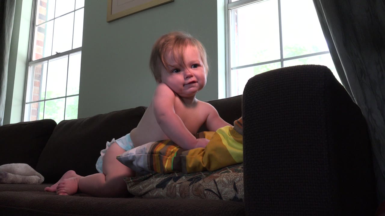 At 0:00, a baby in a diaper sits on a dark couch in a Westminster, Colorado home, with pillows beneath them and a bright window behind.