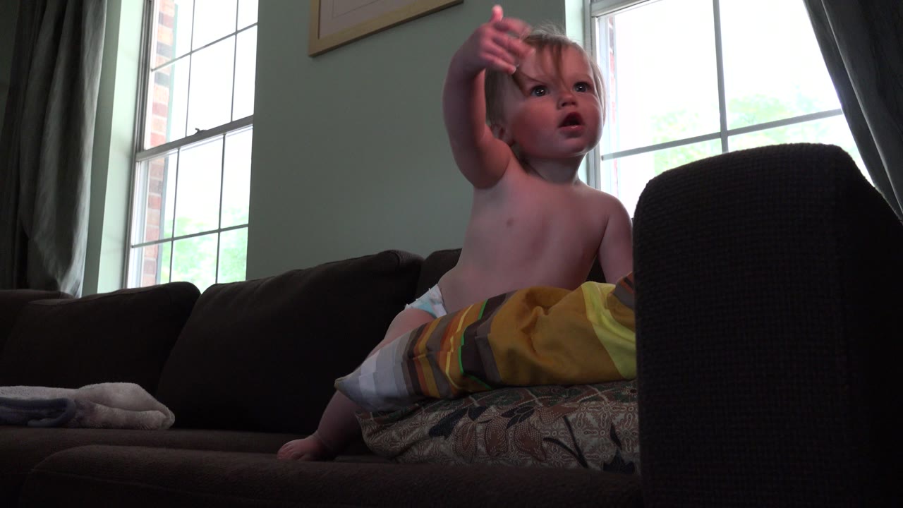 Small child sitting on a couch and pointing upward near a bright window.