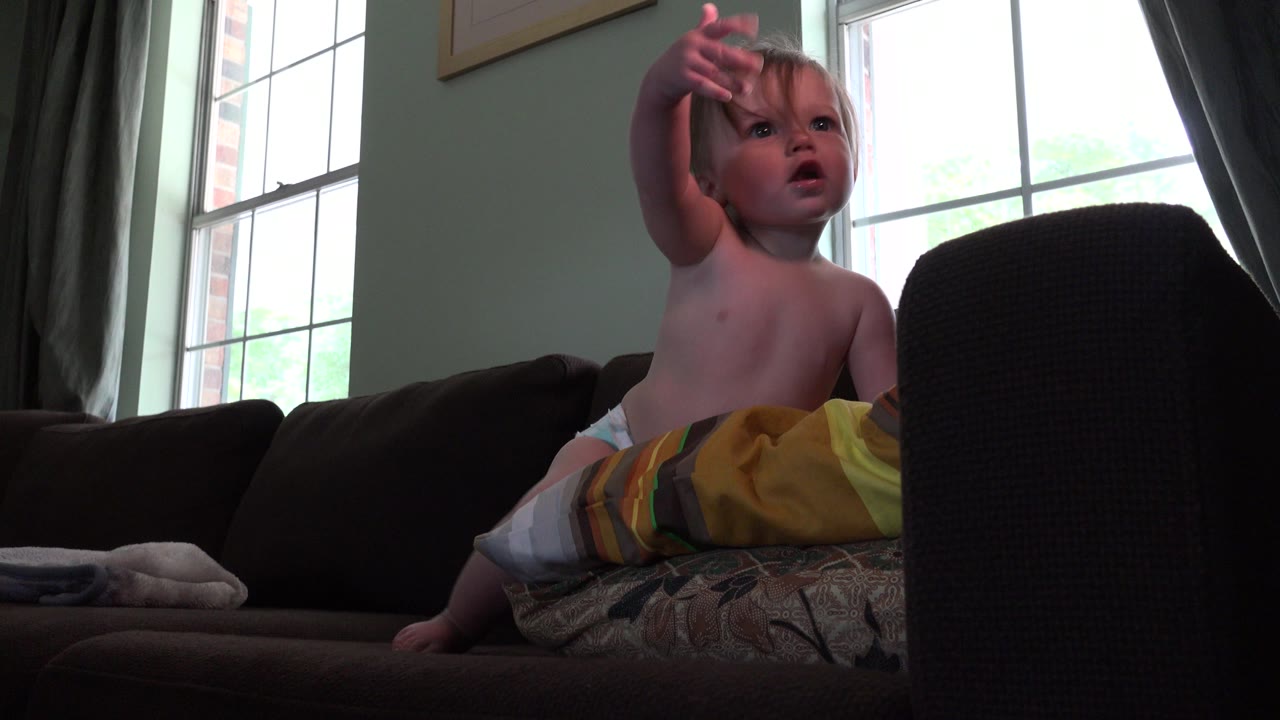 The child remains seated on the couch in the Westminster, Colorado interior, raising a hand while the window light outlines the couch arm and curtains.