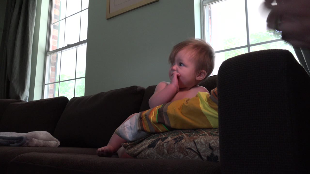 A toddler sits on a dark couch in a Westminster, Colorado home while a bright window backlights the room and part of an adult face appears near the right edge.