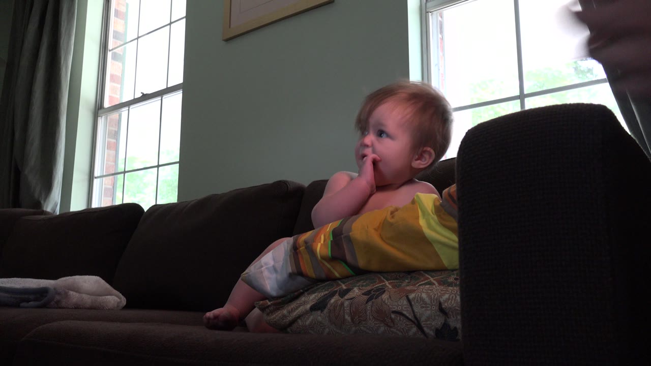 The toddler reclines on the couch in the Westminster, Colorado living room, with fingers near the mouth and a partially visible adult face at the right edge.