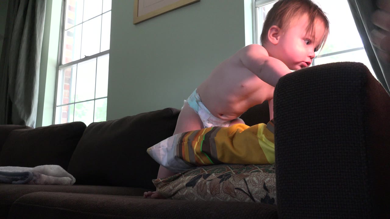 Blond baby in a diaper leaning on pillows on a dark sofa.