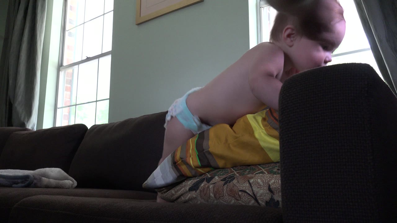 Baby in a diaper leaning over pillows on a sofa near a chair.