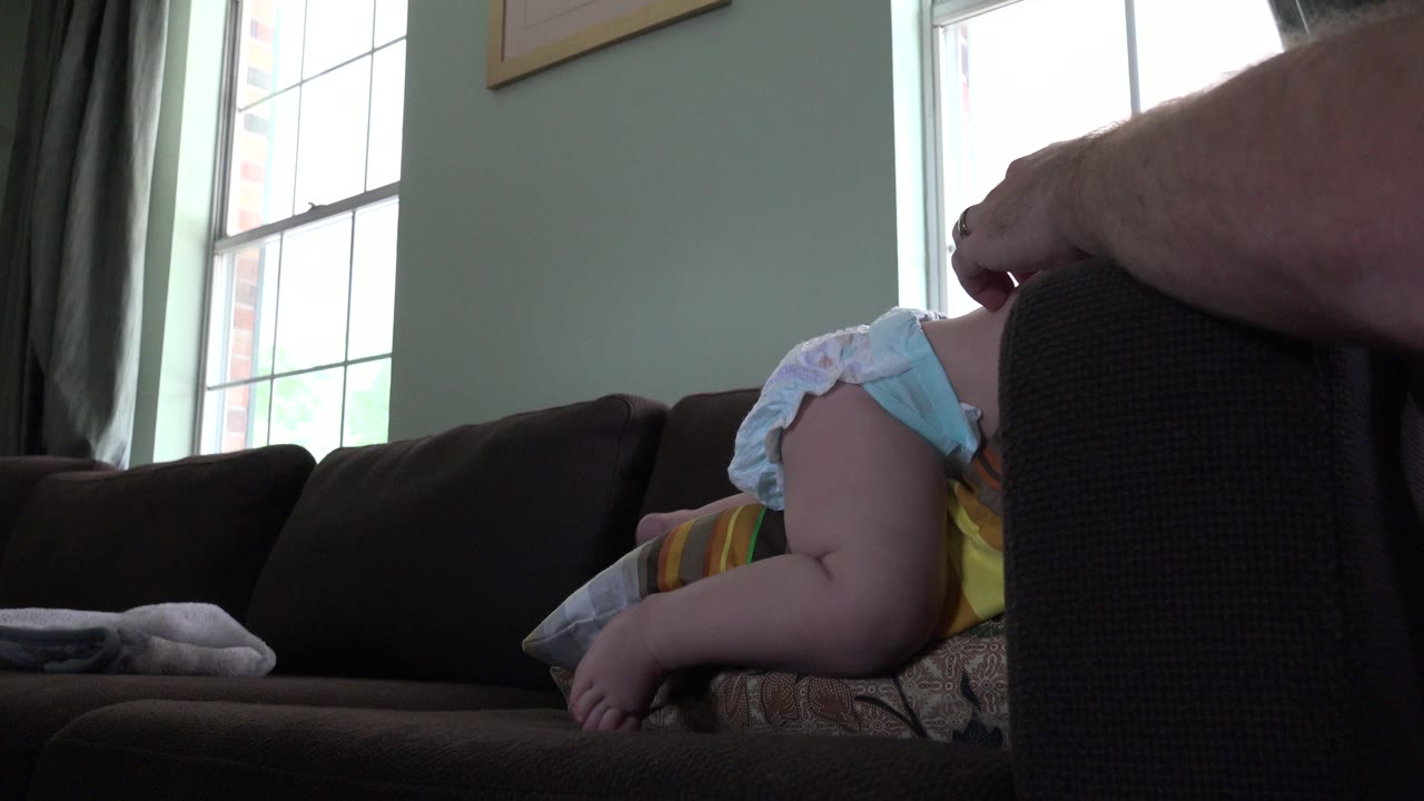 Young child partly upside down on a brown sofa near bright windows.