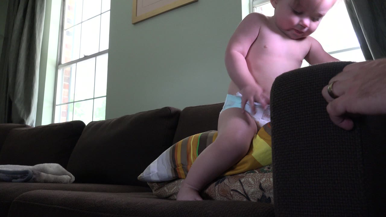 Baby in a diaper sitting on a dark sofa near an adult hand.