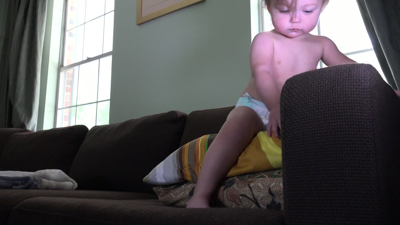 Toddler in a diaper stands on a sofa behind a dark cushion.