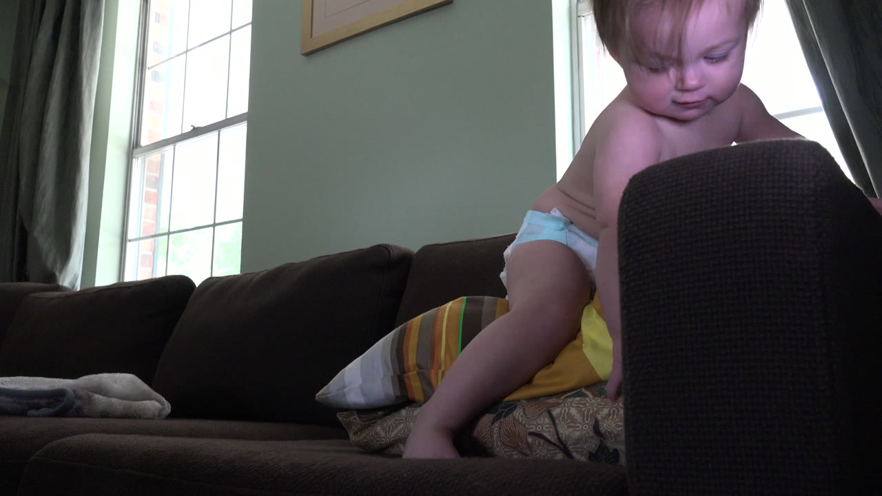 Toddler in a diaper standing on a sofa near a dark chair back.