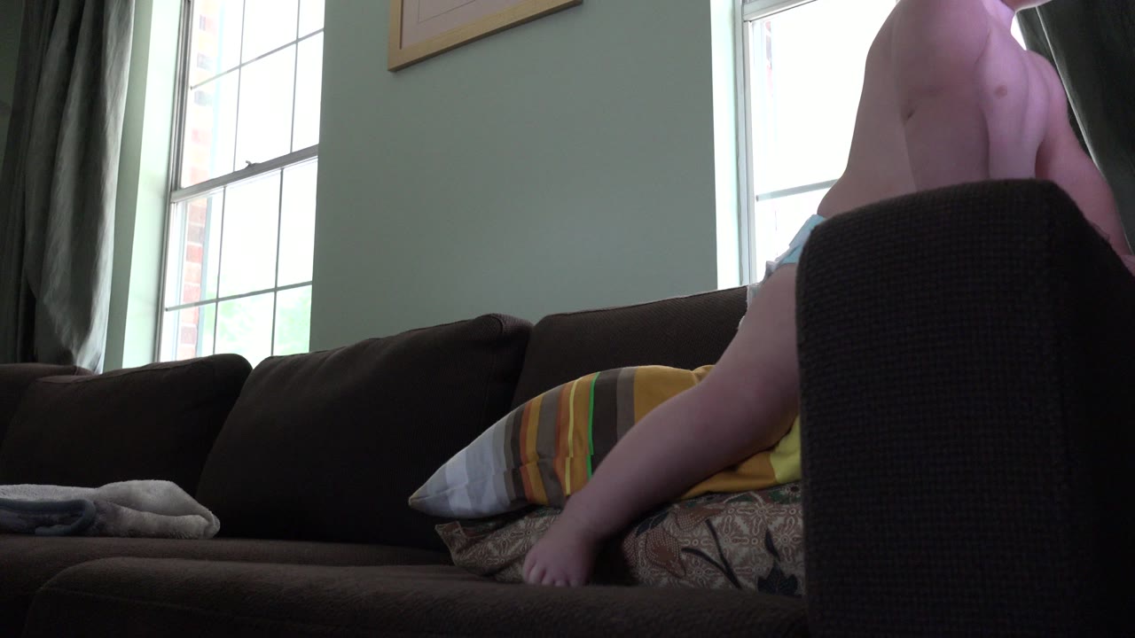 At 0:01, the baby is still upright beside the sofa, with one leg visible near striped pillows and the tall window backlighting the room.