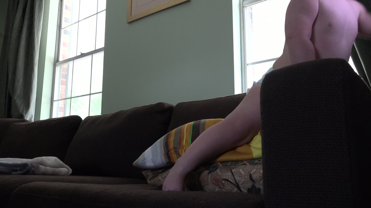 Diapered child standing on a couch near bright windows.