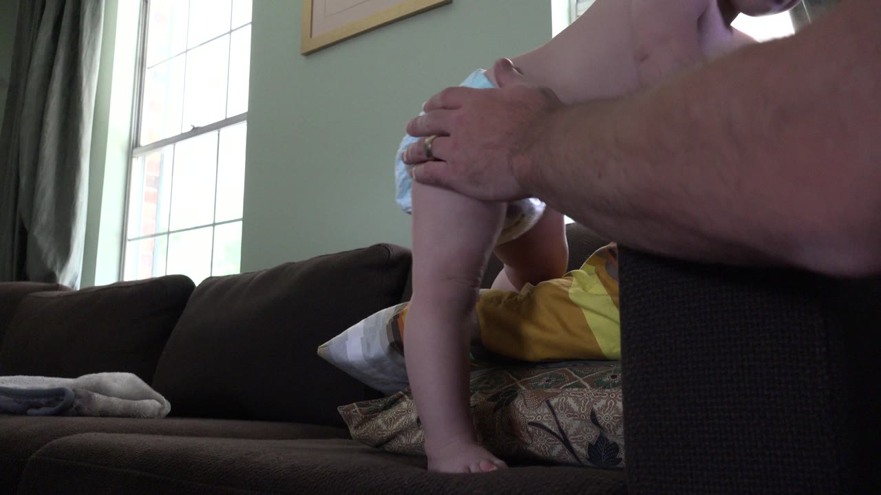 A close low view shows the adult hand on the baby's back while the child braces on the couch in the Westminster, Colorado home interior.