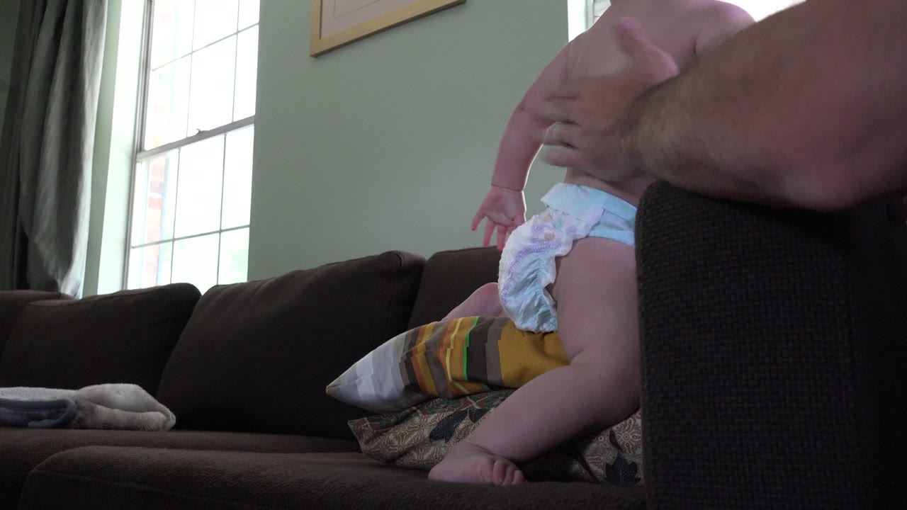 The baby remains upright on the couch in the Westminster home, with the adult hand visible across the upper right and pillows stacked behind the child.