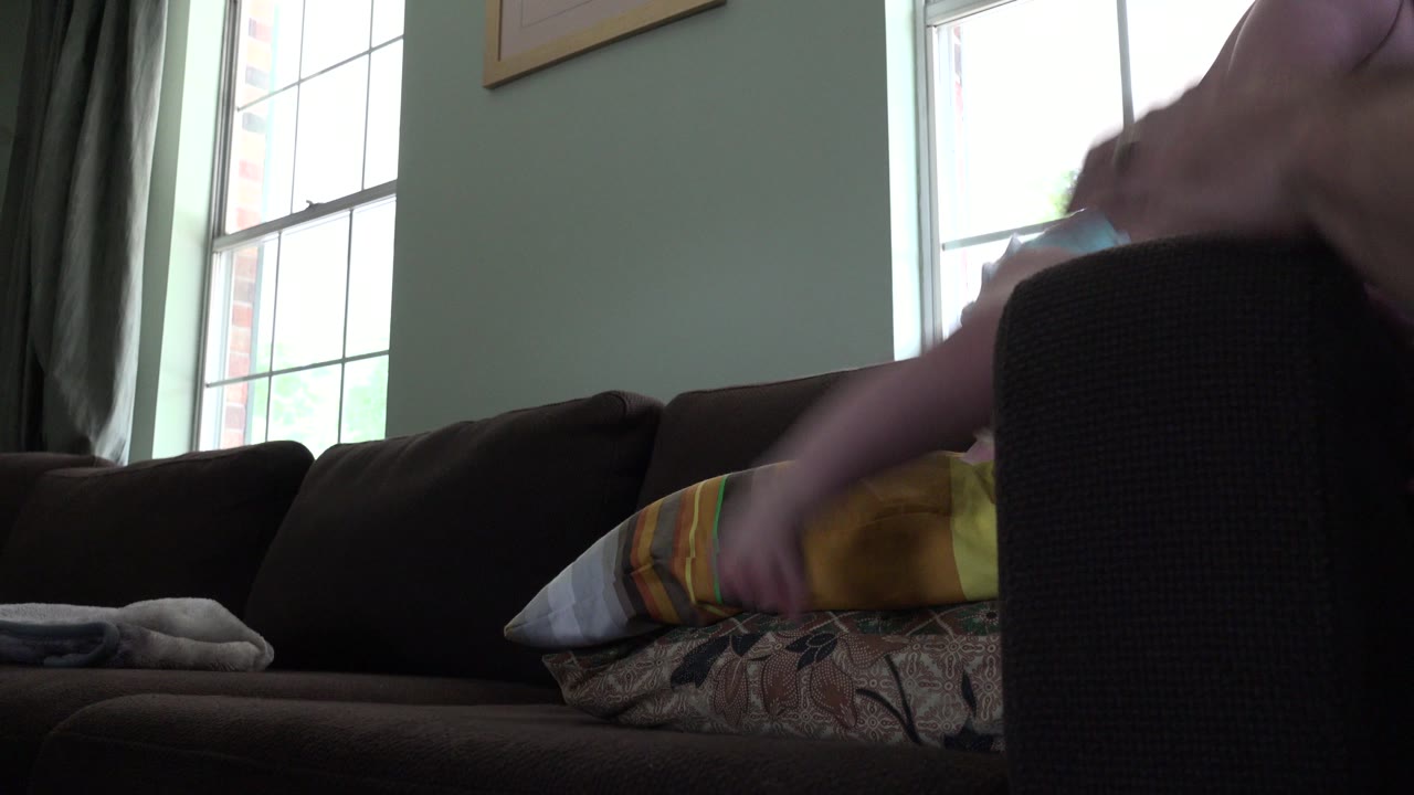 In Westminster, Colorado, the baby is partly obscured by the dark sofa arm while one arm reaches toward pillows on the couch in the backlit living room.