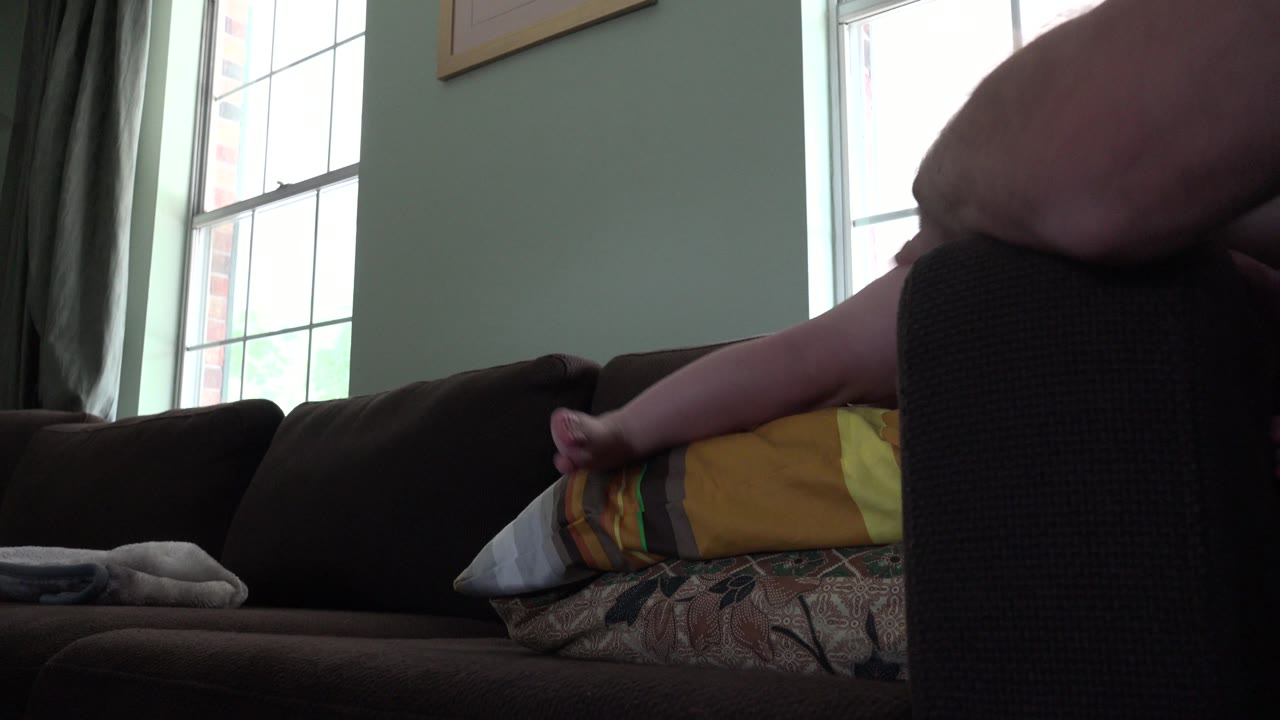 Baby's leg and foot rest near pillows on a sofa.