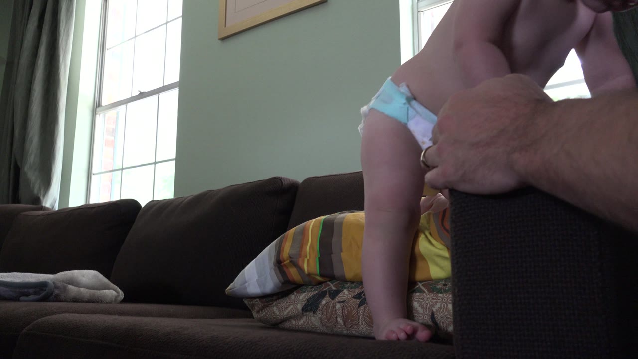 In the Westminster home interior, the toddler shifts position on the pillows, with one foot lower near the couch cushion and the adult arm still close.