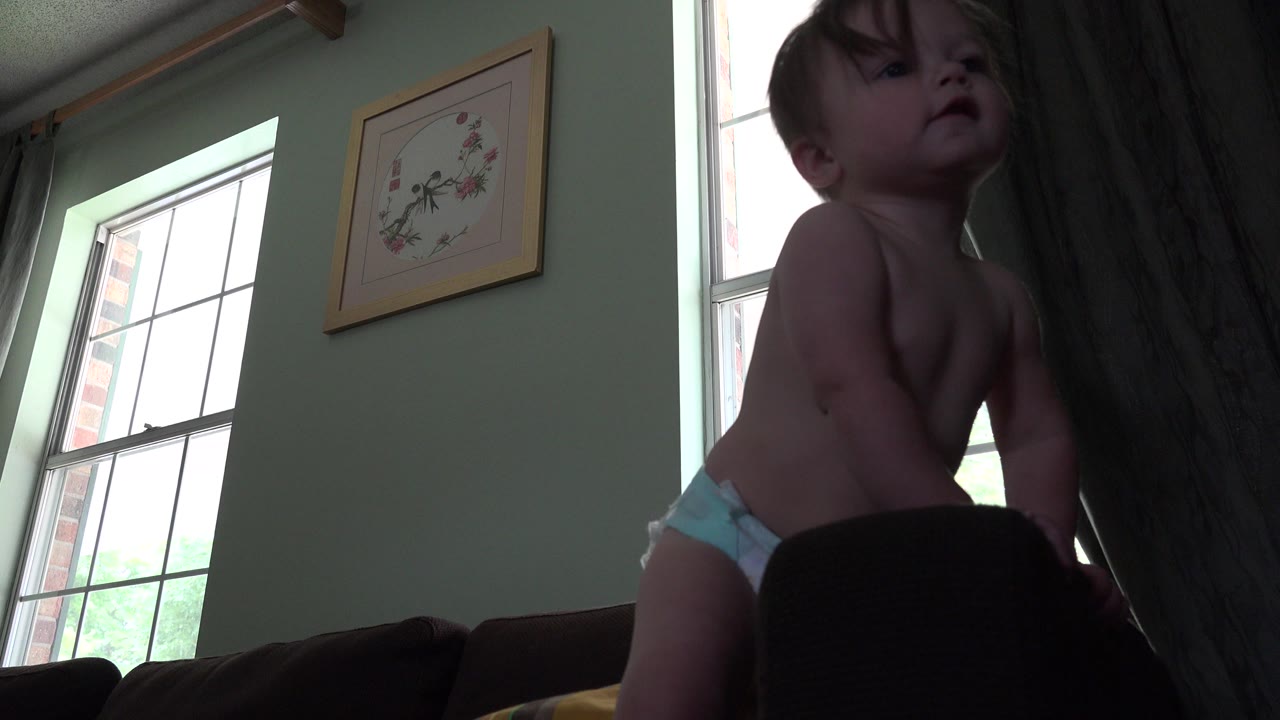 Baby in a diaper leaning on dark furniture beside bright windows.
